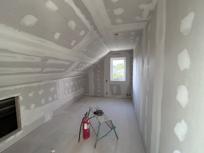 Nymålat rum under renovering med spacklade väggar och tak, fönster i änden av rummet, en stege och brandsläckare i förgrunden.