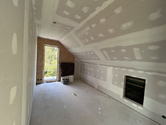 Rum under renovering med spacklade gipsväggar, sluttande tak och en stor fönsteröppning med utsikt över grönska.