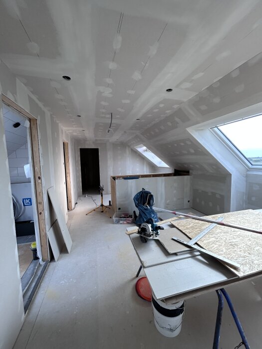 Rum under renovering med spacklade väggar och tak, byggutrustning på ett bord och öppna dörrar. Ett stort fönster ger ljusinsläpp.
