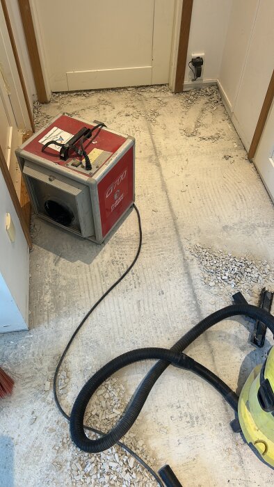 En industridammsugare och sladdar på ett delvist borttaget klinkergolv med damm och skräp.