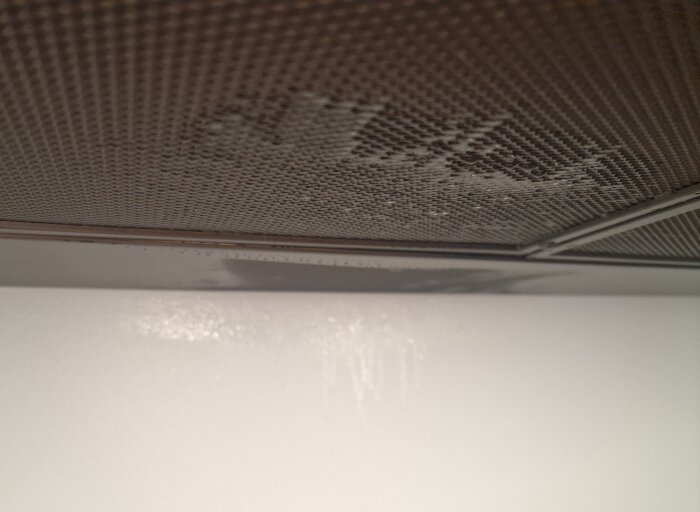 Närbild av ett köksfläktfettfilter där vatten eller kondensdroppar syns vilket har orsakat blöta fläckar på väggen nedanför.