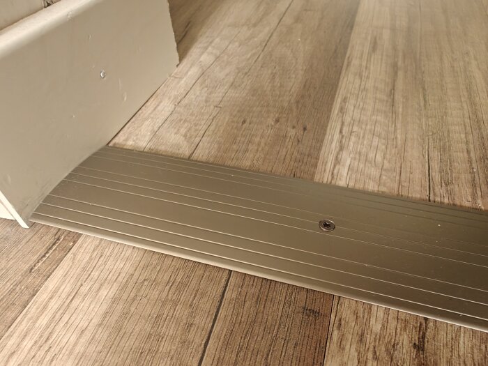 Tröskelplatta av metall fastskruvad på trägolv mellan två rum.