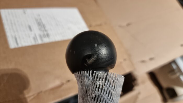 Bild av en dragkrok i plastförpackning med repor och skador på ytan, ovanpå en kartong.