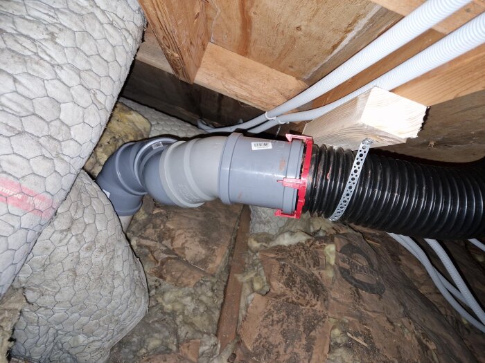 Grå ventilationsslang ansluten på vinden, förstärkt med extra regel mellan träkonstruktion, med isolering och plywood synligt.