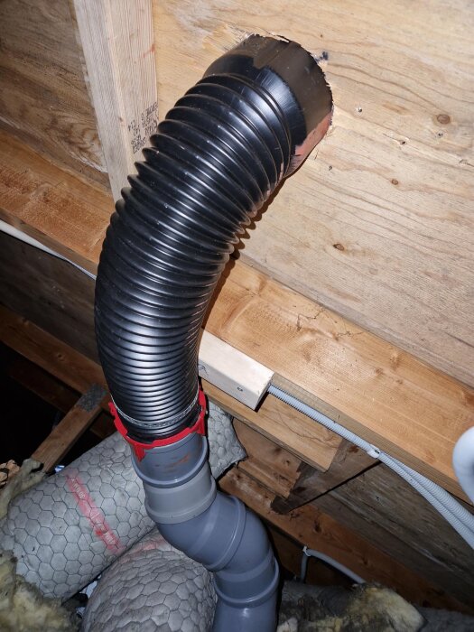 Flexibel svart slang installerad på vind, fäst genom plywood och mot takstolar med en extra regel. Synlig isolering och ett grått rör under.