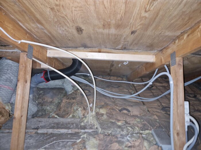 Isolerad vind med ny slangdragning, extra regel mellan takstolar och hängande plywood synligt, tillsammans med diverse kablar och isoleringsmaterial.