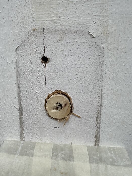 Hål i vitmålad fasadpanel med sprickor runt ett tidigare vattenutkast. Maskeringstejp täcker nedre delen av bilden.