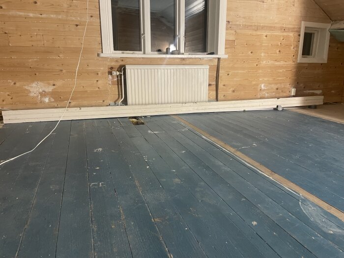 Lutande trägolv i sekelskifteshus med blåa plankor. Rummet har en vägg med träpanel, ett fönster och ett element.