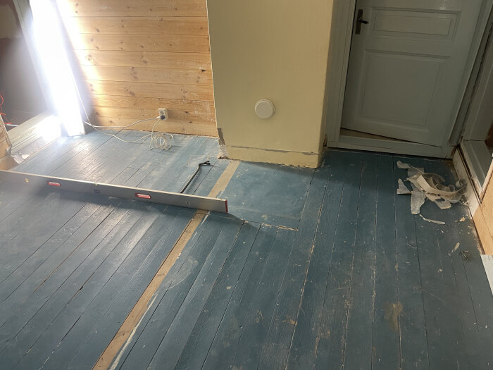 Blåmålat trägolv i äldre sovrum med vattenpass som visar golvets lutning, dörr i bakgrunden och delar av väggen träbeklädd.