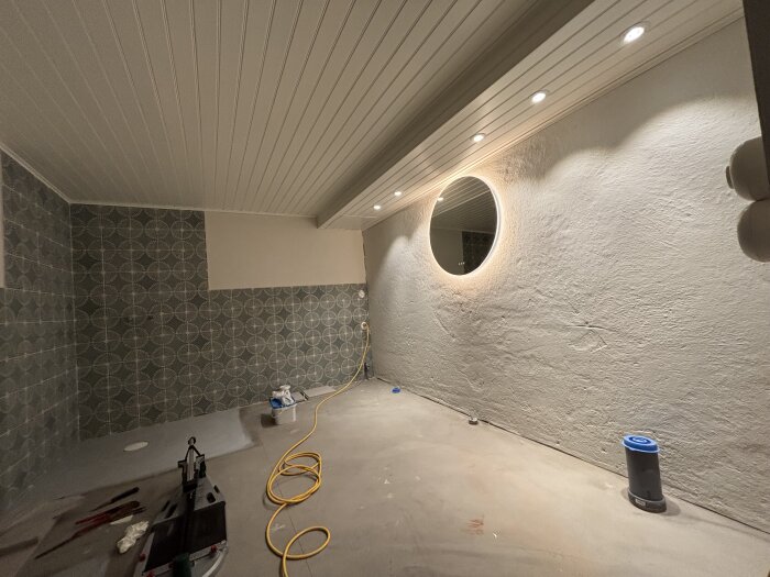 Renoverat rum med paneltak, infällda lampor, runt upplyst spegel på råputsad vägg, mönstrad kakel, kablar och verktyg på golvet.