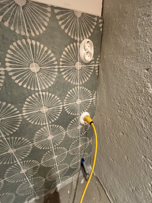 Vägg med dekorativa kakelplattor, ett dubbeluttag och en gul elkabel.