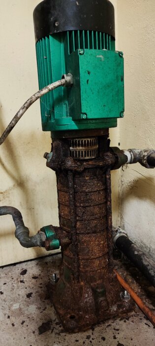 Äldre Grundfos pump med rost, röranslutningar och elektrisk ledning, installerad mot en vägg.