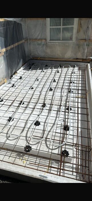 Bild av en byggarbetsplats med golvvärmesystem under installation, inklusive rör lagda på armeringsnät intill en plasttäckt vägg och fönster.