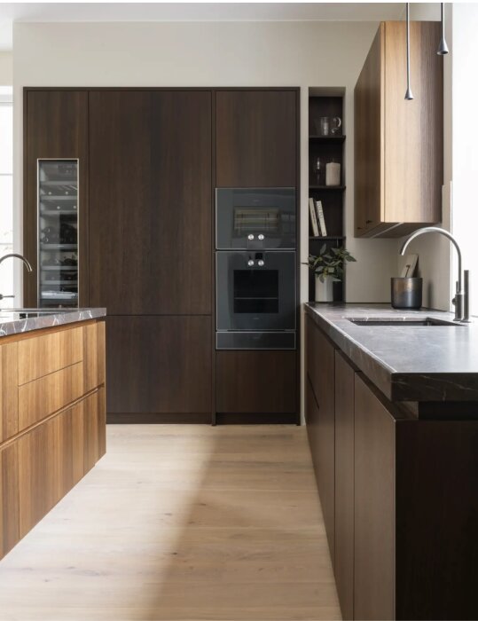 Kök med mörka träskåp, vinkyl och dubbelugn, ljust trägolv och moderna bänkskivor i marmor, samt integrerade hushållsapparater.