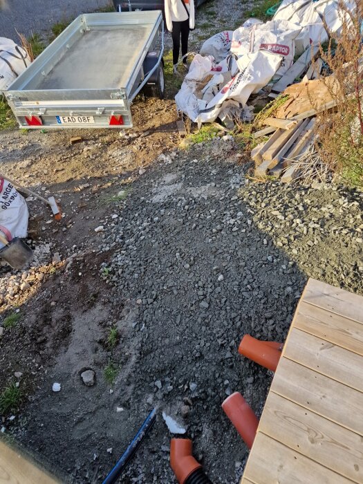 Grusfylld ytavlastning med orangea dräneringsrör, omgiven av påsar samt en släpvagn i bakgrunden.