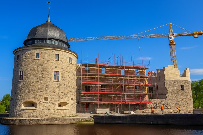 Ett stenslott under renovering med byggställningar och en byggkran. Arbetare syns arbeta vid slottets murar vid vattnet.
