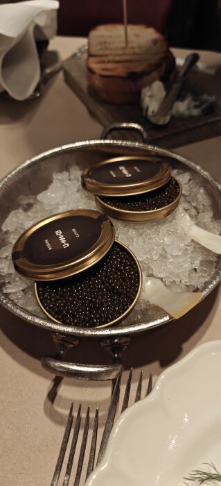 Två burkar svart caviar på is i en metallskål, serverade med smörgåsar och kräm.