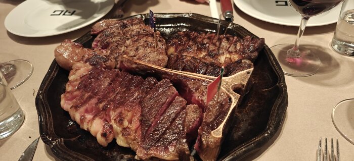 En tallrik med grillade köttbitar, vissa med ben, omgiven av vinglas på en restaurang med AG-logotyp.