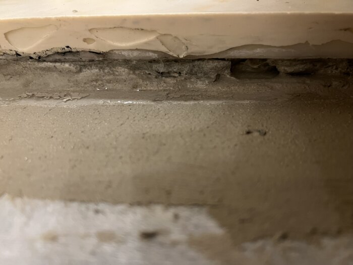 Skadat golv under tröskel med blött tätskikt och ojämna ytor, del av byggprojekt där fuktproblem åtgärdas med avfuktare.