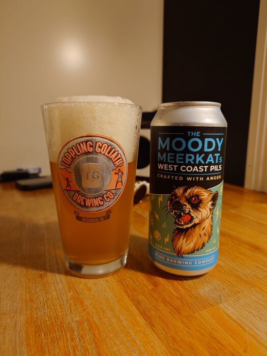 Ölglas med skummande öl från Toppling Goliath bredvid en burk "The Moody Meerkat's West Coast Pils" på ett träbord.