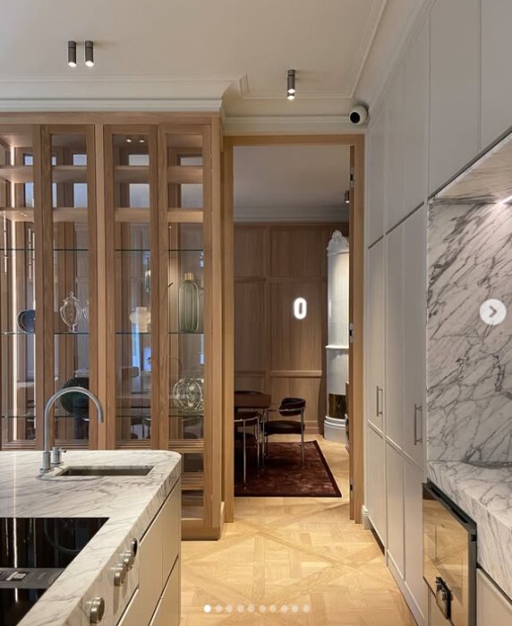 Modern köksinteriör med ljusa skåp, marmorbänkskiva och öppen hylla med glas. Vy mot matplats i bakgrunden.