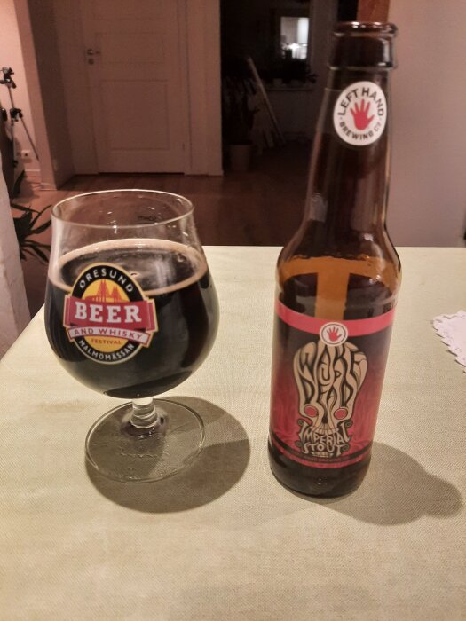 Glas med mörk öl bredvid flaska med etikett "Wake Up Dead Imperial Stout" från Left Hand Brewing Co. på bord.
