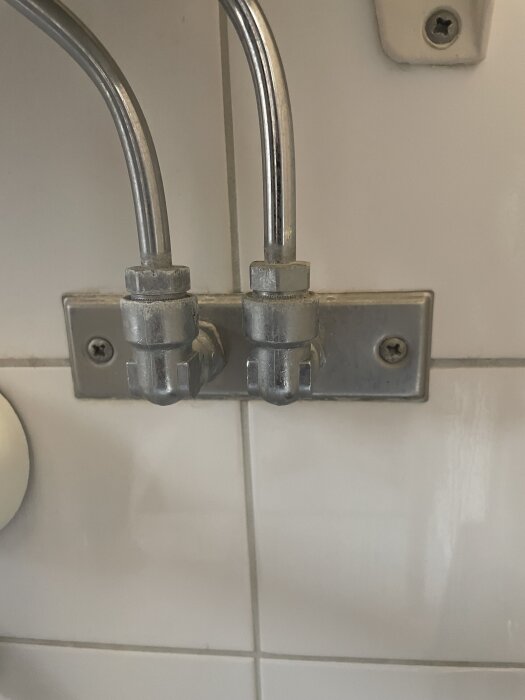 Två metallkopplingar med rör på en vit kakelvägg under ett handfat.