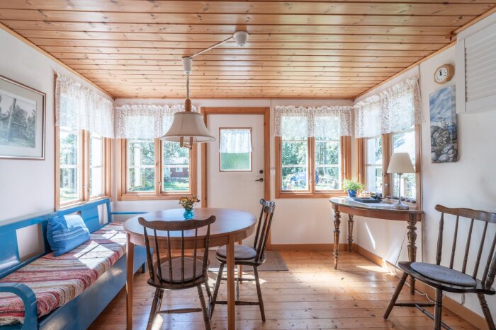 Ljust rum i sommarhus med trägolv, träfönster och möbler i rustik stil. Solen skiner in genom spetsgardiner över fönsterbänken.