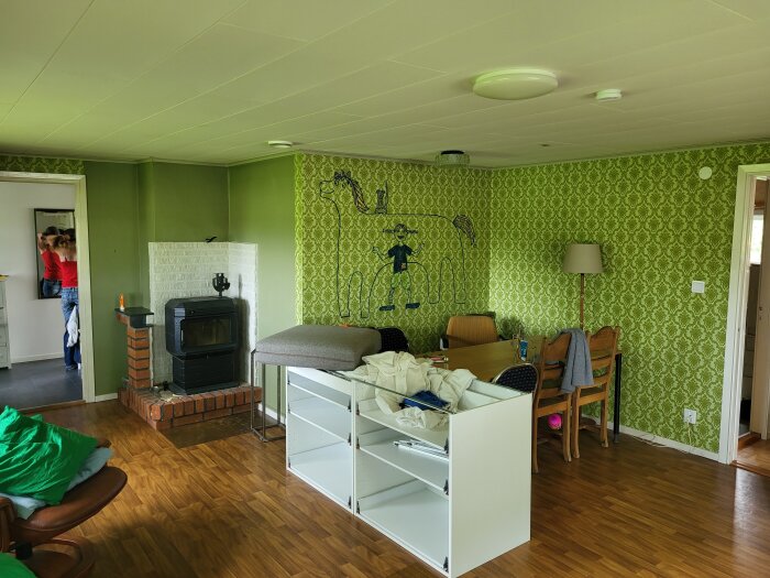 Grönt vardagsrum med en vit öppen spis och kamin. En vit byrå, gröna väggar med tapet och ett träbord mot väggen med stolar runt.