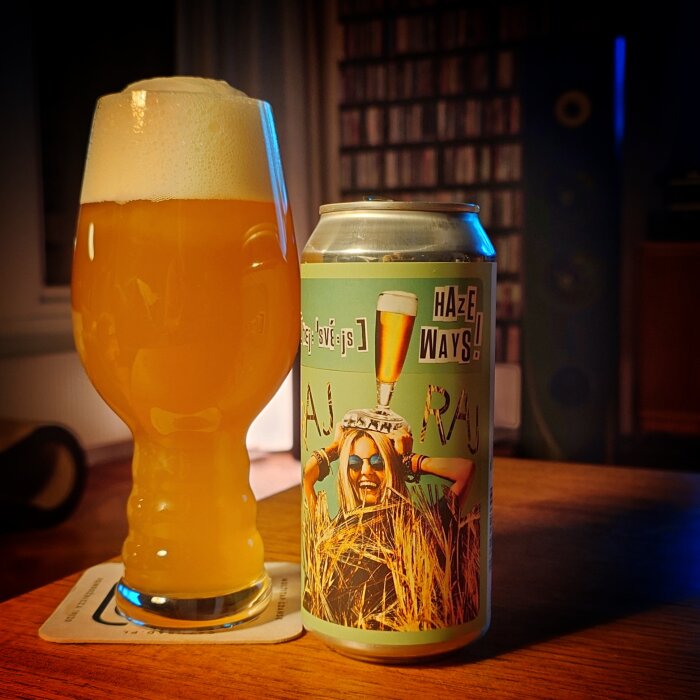 Ölglas med skummande öl och en ölburk bredvid, märkt "Haze Ways", på ett bord i ett rum med dämpad belysning.