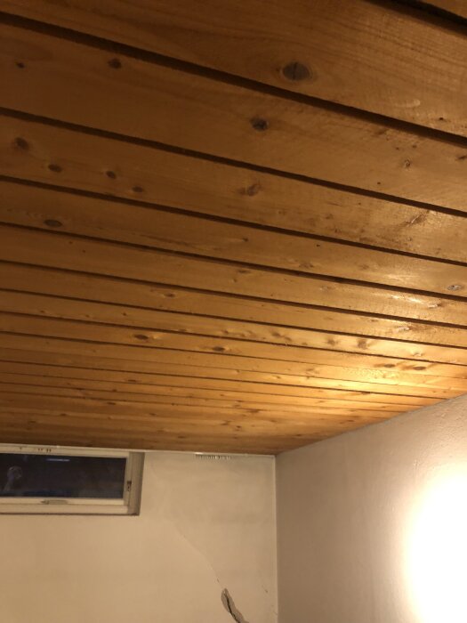 Trätak i källare med panelbrädor, gediget hantverk från 1960-talet. Fönster och spricka syns på väggen under taket.