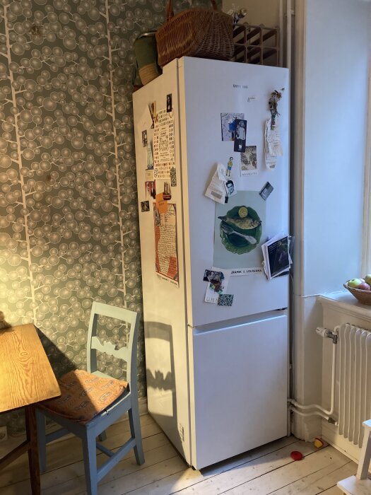 Kylskåp i kökshörn med dekor på dörren, bredvid en blå stol och ett trämönstrat tapetbord, förvaringskorg ovanpå.