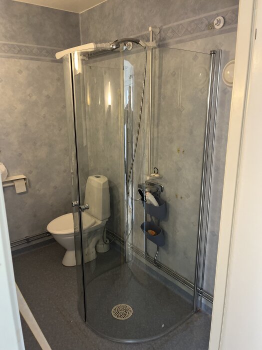 Duschhörna med glasdörrar i ett badrum, toalettstol bredvid, blå väggar, monterade duschdörrar som behövde bytas på grund av fabrikationsfel.