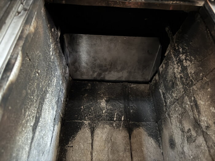 Rökgashylla i en kamin med sot- och smutsspår, nyinstallerad plåt ersätter den gamla, visar insidan före installation av lufttätningslist.