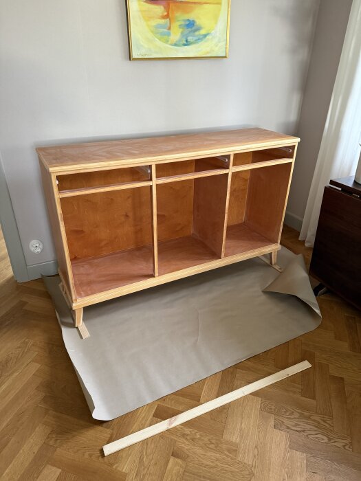 Vitrinskåp utan dörrar och beslag under renovering på ett parkettgolv, med skyddspapper och en trälist framför.