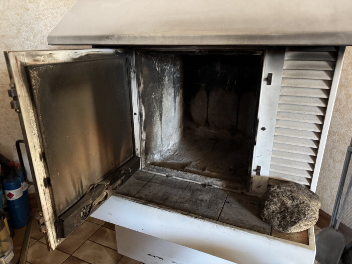 Öppen kamin med dörren på glänt, insidan sotig och smutsig. En sten ligger på kanten av kaminen. En rökgashylla nämns i texten.