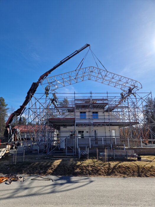 En kran lyfter en takstol över ett hus med byggnadsställningar, under en dag med blå himmel.