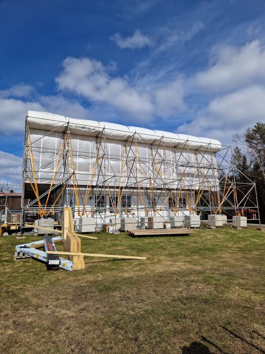 Byggnadsställningar omger ett hus med en överbyggnad av plast för skydd under takrenovering. Grön gräsmatta i förgrunden.
