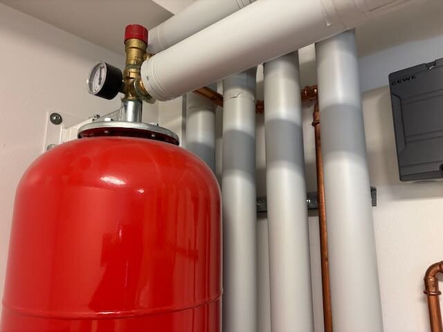 Röd expansionstank med ett tryckmätare omgiven av flera vita och kopparfärgade rör.