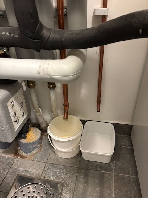 Dräneringsrör från varmvattenberedare och expansionstank med plastlådor för att samla upp droppande vatten på ett golv med golvbrunn.