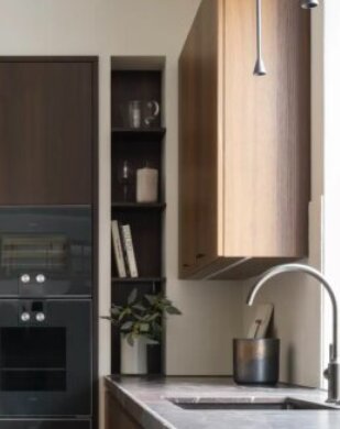 Köksinteriör med diskho, mörka skåp, inbyggd ugn, öppna hyllor med böcker och dekorativa föremål, och en kran över bänkskivan.
