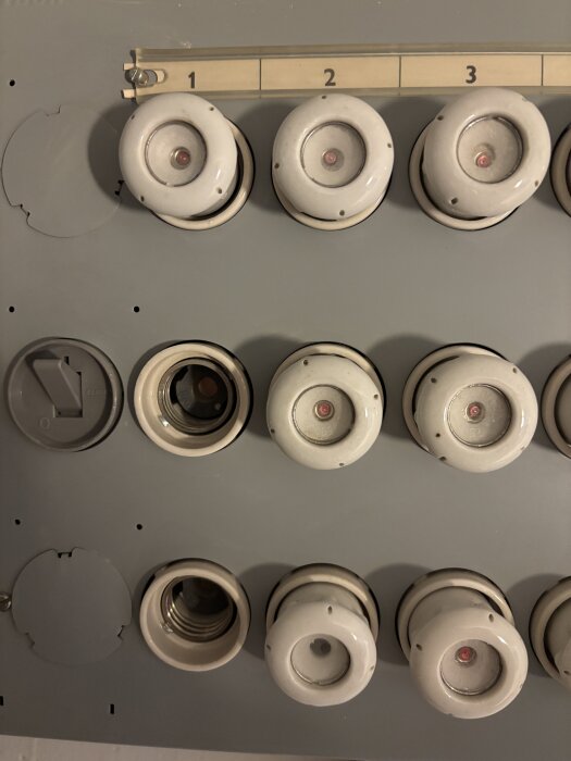 Grå hållare med flera vita proppsäkringar ordnade i rader, märken för olika nummer syns ovanför proppsäkringarna.