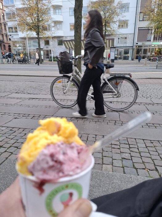 En hand håller en bägare med tvåsorters glass, en person går förbi cyklande i stadsmiljö.