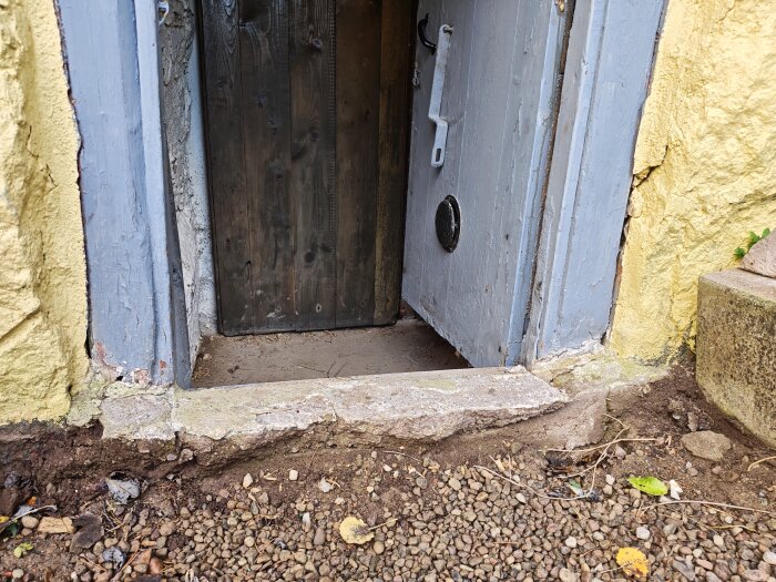 Bild av en källaringång, synlig dörr med blått trä och omgiven av gul puts. Grus och jord framför ingången utan trappsteg.