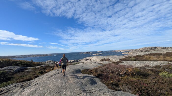 En person promenerar på klippor vid kusten under en klarblå himmel, med havet och flera små öar i bakgrunden.