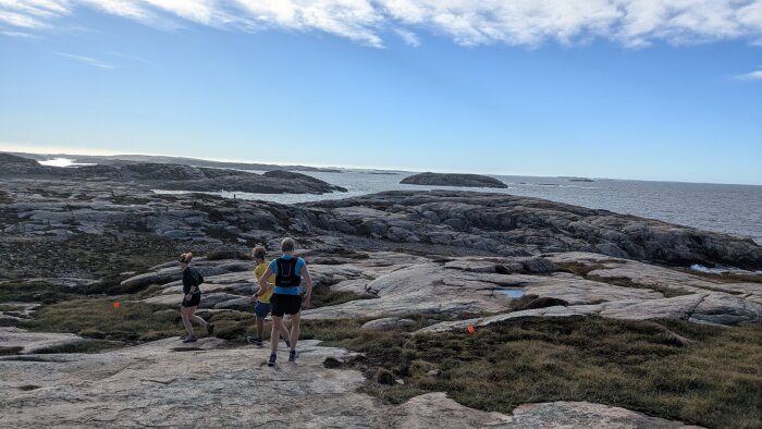 Tre personer vandrar på klippor med havsutsikt under en klarblå himmel.