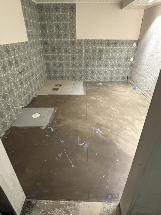 Del av badrum under renovering med nylagd kuvertskärning på golvet. Väggar med mönstrat kakel, resten av golvet är primat.