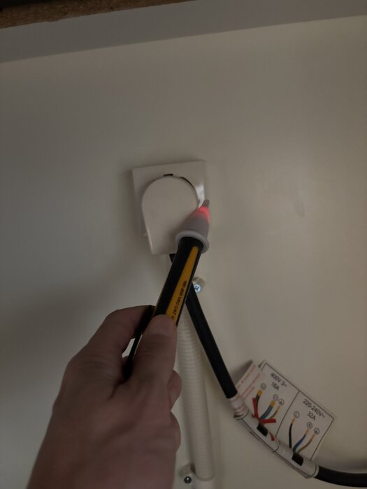 Hand som håller en spänningstestarpenna vid en spisanslutning, med en lampa som lyser vid den högra sidan av uttaget.