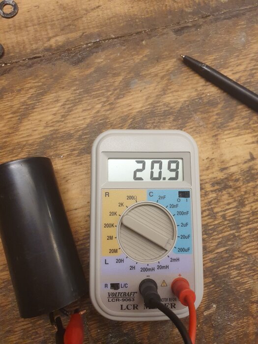 Multimeter visar 20,9uF, bredvid en svart kondensator, på ett träbord.