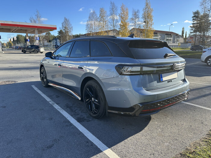 Grå Volkswagen GTX parkerad vid en bensinstation under en klarblå himmel, omgiven av träd och byggnader i bakgrunden.
